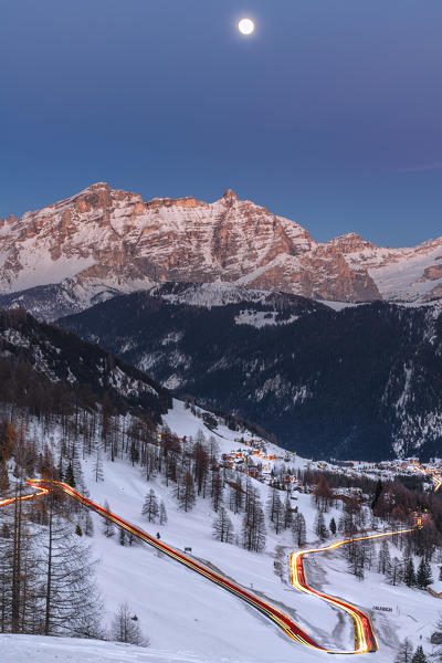 Colfosco, Alta Badia, province of Bolzano, South Tyrol, Italy, Europe. Car lights on the Gardena pass road. In the background the lights of the villages Colfosco and Corvara, the peaks of Lavarella and Conturines and the full moon