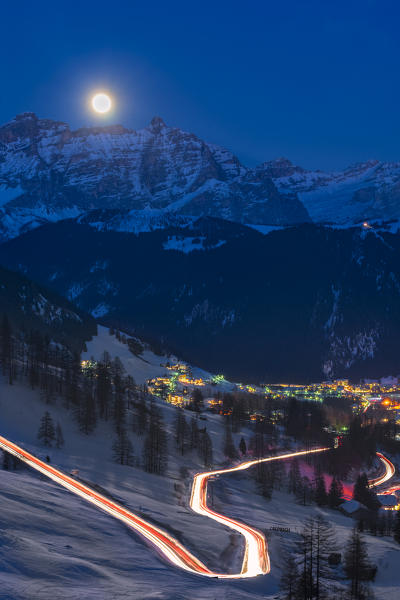 Colfosco, Alta Badia, province of Bolzano, South Tyrol, Italy, Europe. Car lights on the Gardena pass road. In the background the lights of the villages Colfosco and Corvara, the peaks of Lavarella and Conturines and the full moon