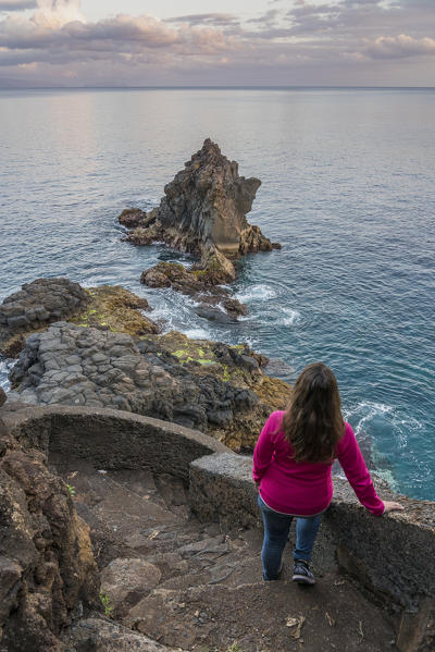 Woman observing the Atlantic Ocean and rock formations at dusk. Santa Cruz, Madeira region, Portugal.