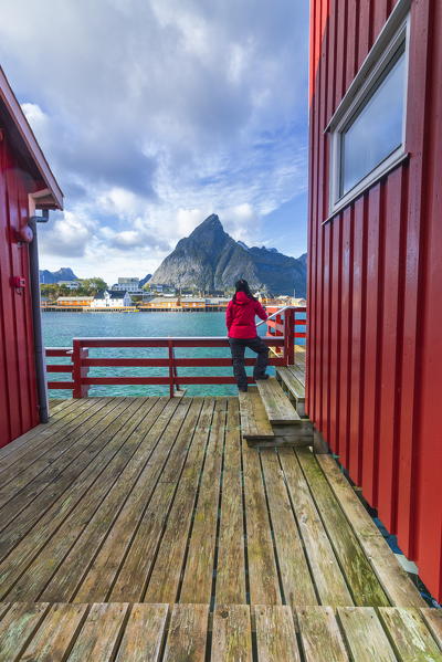 Woman on wooden deck look towards Sakrisoy and  Olstinden mountain, Reine, Nordland, Lofoten Islands, Norway