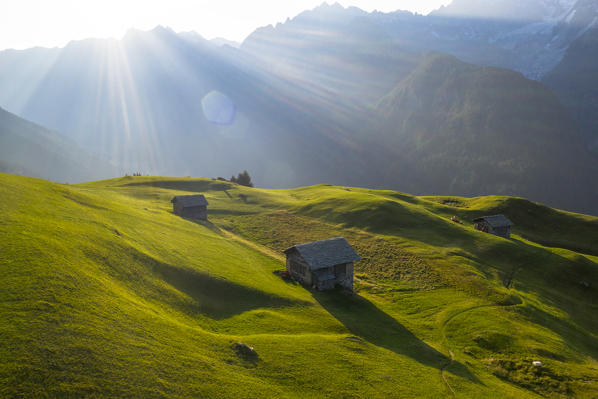 Aerial view of huts in the green meadows of Tombal Alp lit by sun rays, Soglio, Val Bregaglia, canton of Graubunden, Switzerland