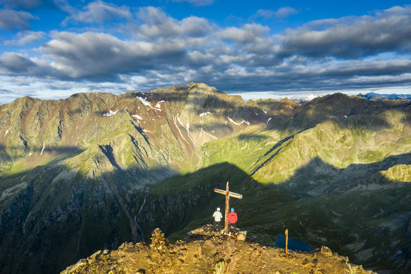 Two hikers admiring landscape from the summit cross on Monte Gaviola, aerial view, Gavia Pass, Valfurva, Valtellina, Lombardy, Italy