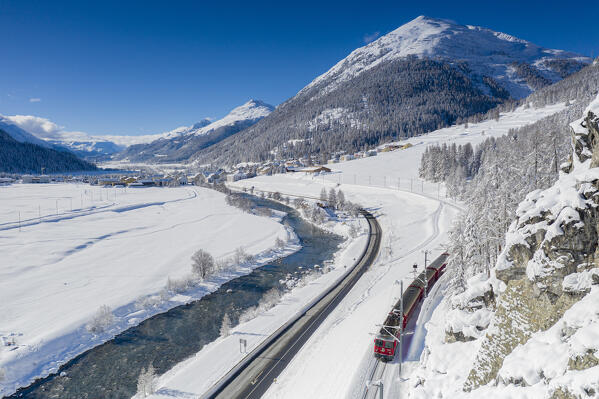 Bernina Express train traveling in the snowy landscape on shore of Inn river, Madulain, Graubunden, Engadin, Switzerland