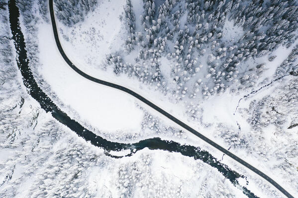 Aerial view of empty mountain road running beside a frozen river in the winter forest covered with snow