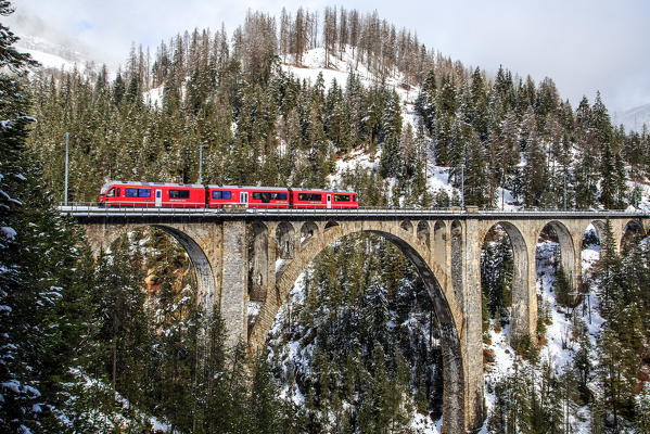 The red train on the Wiesener Viadukt surrounded by snowy mountains and woods. Engadine. Canton of Graubuenden. Switzerland. Europe.