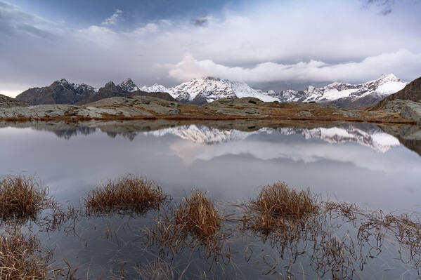 Foggy sky over the snowy mountains mirrored in water in autumn, Alpe Fora, Valmalenco, Valtellina, Lombardy, Italy