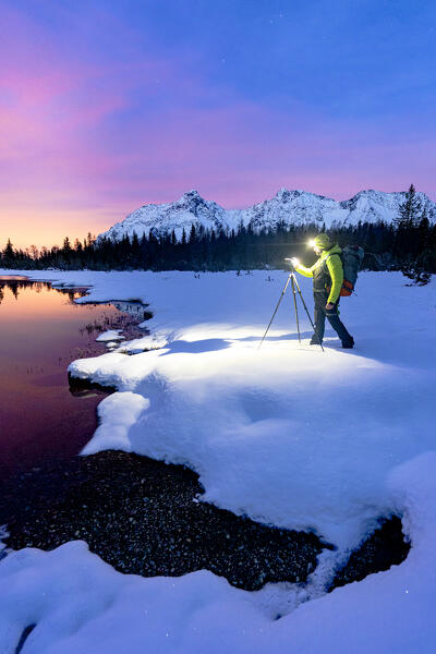 Man with tripod photographing lake Entova at dawn standing on the snowy shores, Valmalenco, Valtellina, Lombardy, Italy