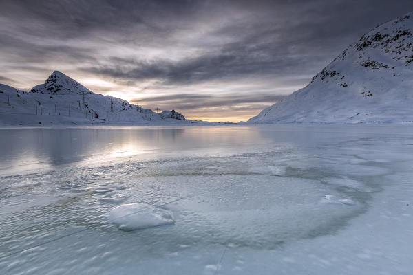 The smooth ice and choppy splits are enhanced by a dark sky clouded by fanciful glazes. White Lake. Bernina Pass. Canton of Graubuenden. Engadine. Switzerland. Europe