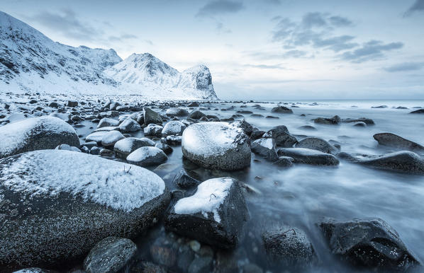 Rocks in the cold sea and snow capped mountains under the blue light of dusk Unstad Lofoten Islands Northern Norway Europe