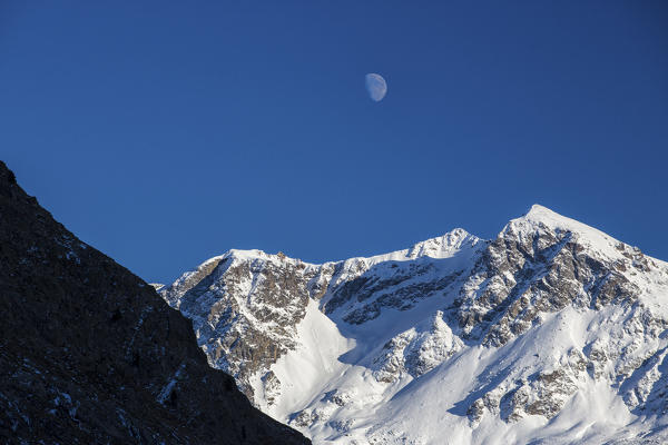 The moon appears above the snowy peaks in the blue sky Julierpass Albula District Canton of Graubünden Switzerland Europe