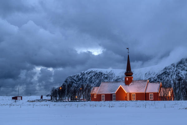 Lights on the church at dusk with the snowy peaks in the background Flakstad Lofoten Islands Norway Europe