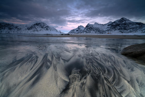 Waves and ice on the surreal Skagsanden beach surrounded by snowy peaks Flakstad Nordland county Lofoten Islands Norway Europe