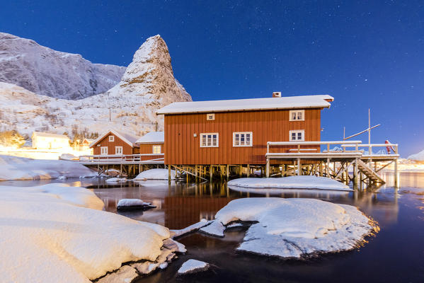 Starry night on the snowy peaks surrounded by the frozen sea and fishermen houses Reinevagen Bay Lofoten Islands Norway Europe