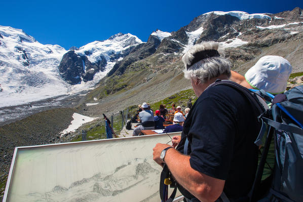 Hikers reading the names of the snow-capped peaks of the Bernina group from a map on the terrace of the Chamanna Boval Hut - Engadine, Switzerland Europe