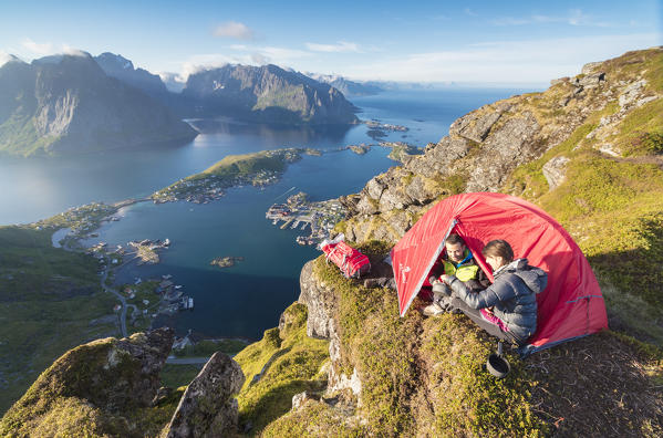 Hikers in a tent on the top of peak admire the blue sea framing the village Reinebringen Moskenes Lofoten Islands Norway Europe