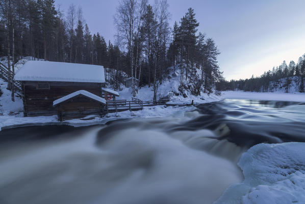 A wooden hut surrounded by the river rapids and snowy woods at dusk Juuma Myllykoski Lapland region Finland Europe