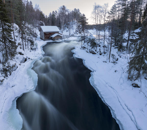 A wooden hut surrounded by the river rapids and snowy woods at dusk Juuma Myllykoski Lapland region Finland Europe