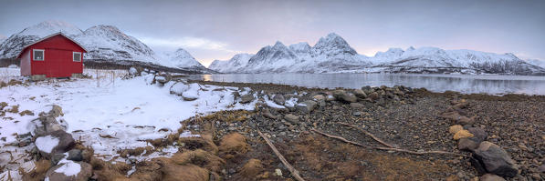 Panorama of the wooden hut called Rorbu framed by frozen sea and snowy peaks Svensby Lyngen Alps Tromsø Norway Europe