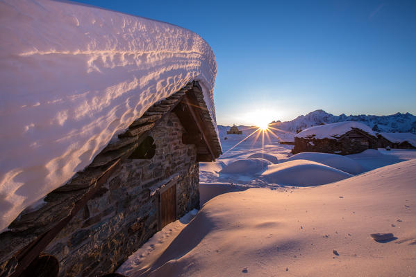 The last rays of sun light up the Prabello alp chalets covered with snow. In the background the silhouette of Mount Disgrazia. Sondrio. Valmalenco. Valtellina Lombardy. Italy. Europe