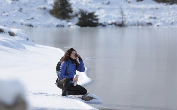 a view of a winter trekking where the woman sits by the lake and drink a cup of tea, Colbricon Lake, Trento province, Trentino Alto Adige, Italy, Europe