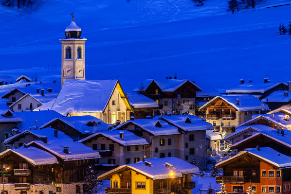 Church and houses illuminated during a winter twilight. Livigno, Valtellina, Lombardy, Italy, Europe.