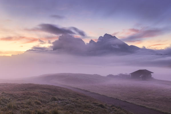 The fog hides a cabin and the Sassolungo group at dawn. Seiser Alm, Dolomites, province of Bolzano, South Tirol, Italy, Europe.