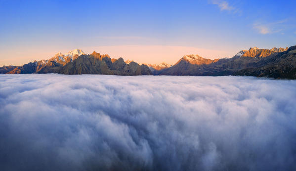 Fog covers the Valmalenco(Val Malenco) with the mountain range of Disgrazia illuminated by sunrise in the background. Valtellina, Lombardy, Italy, Europe.