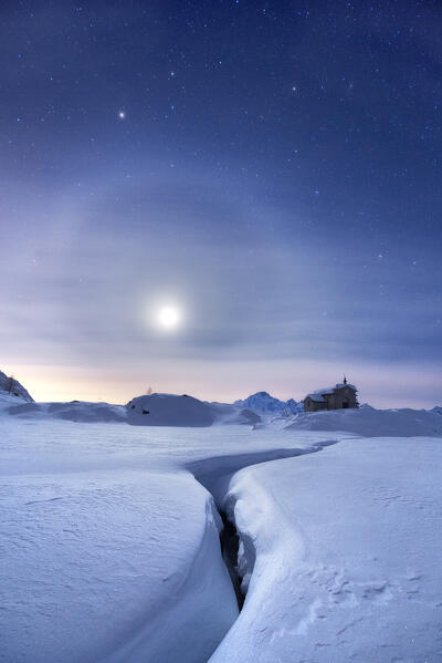 Moon halo in the night sky above a traditional mountain church during a winter full moon night. Valmalenco, Valtellina, Lombardy, Italy, Europe