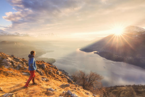 A boy looks the sun rise above the lake. San Bernardo Church, Monte Bregagno, Dongo, Como Lake, Lombardy, Italy, Europe.