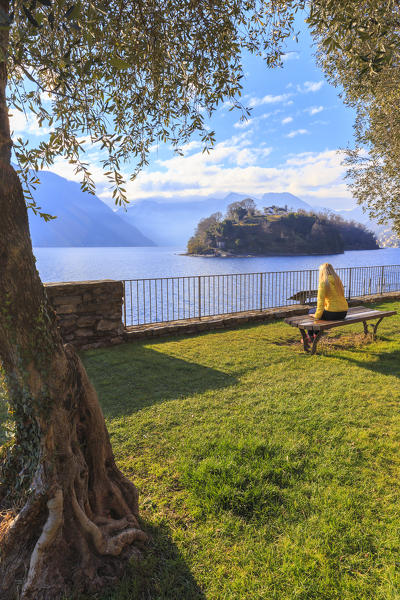 A girl ditting on a bench in front of the Comacina Island, Ossuccio, Como Lake, Lombardy, Italy, Europe.