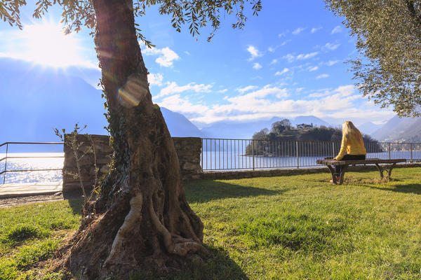 A girl ditting on a bench in front of the Comacina Island, Ossuccio, Como Lake, Lombardy, Italy, Europe.