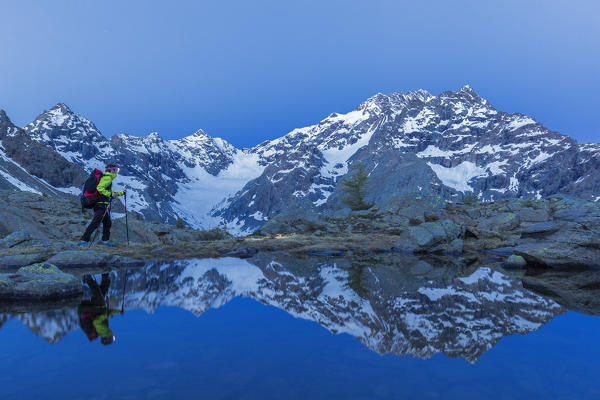 A hiker walks at dusk near a lake that mirrored the mountain range  of Mount Disgrazia. Chiareggio valley, Valmalenco, Valtellina, Lombardy, Italy, Europe.