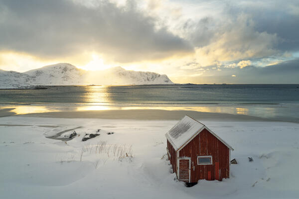 aerial view taken by drone of an classic Rorbuer in to the snow at Ramberg beach during a winter sunset, Ramberg, Lofoten island, Norway, Europe