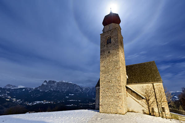 The medieval church of San Nicolò in Monte di Mezzo with the lunar halo. In the background the Dolomite massif of the Catinaccio. Renon plateau, Bolzano province, Trentino Alto-Adige, Italy, Europe.