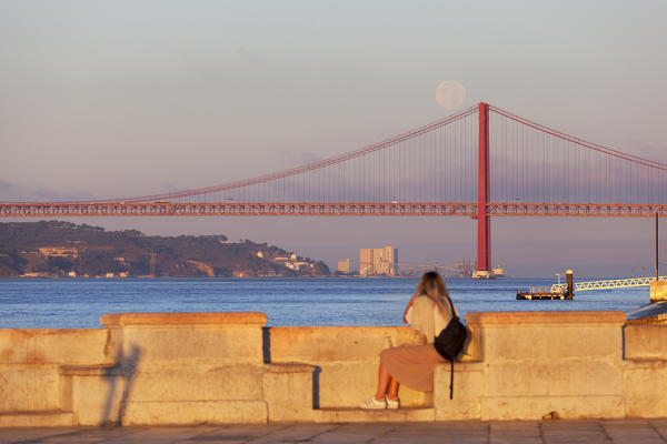 A girl observes the moonset on Ponte 25 de Abril (April 25 Bridge) at dawn, Lisbon, Lisbon Metropolitan Area, Portugal 