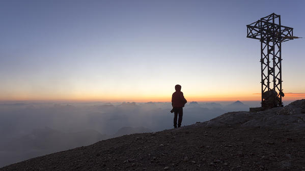 A hiker admires the sea of clouds from the summit cross of Punta Penia, Marmolada group, Dolomites, Canazei, Trento province, Trentino-Alto Adige, Italy