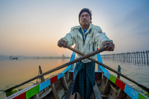 Amarapura, Mandalay region, Myanmar. Man rowing on his colorful boat  on the Taungthaman lake  at sunrise, with the U Bein bridge in the background.