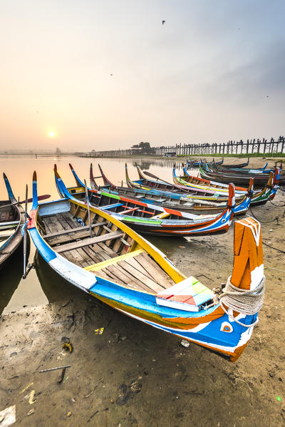 Amarapura, Mandalay region, Myanmar. Colorful boats moored on the banks of the Taungthaman lake  at sunrise, with the U Bein bridge in the background.