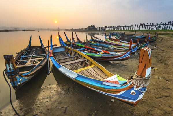 Amarapura, Mandalay region, Myanmar. Colorful boats moored on the banks of the Taungthaman lake  at sunrise, with the U Bein bridge in the background.