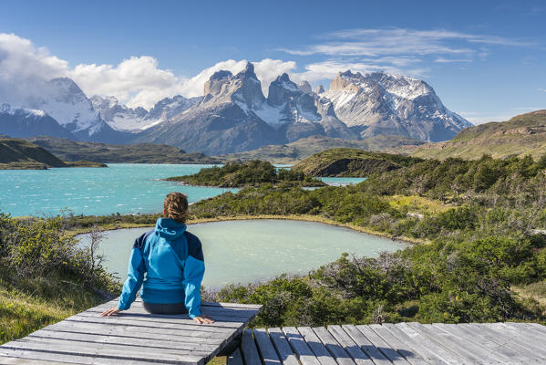 Woman sitting on a boardwalk with Lake Pehoé and Paine Horns in the background, on a windy summer day. Torres del Paine National Park, Ultima Esperanza province, Magallanes region, Chile.