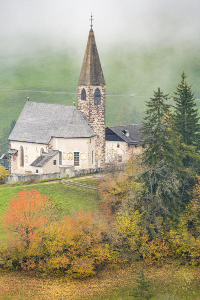 Church surrounded by autumnal trees and mist. Santa Maddalena, Funes, Bolzano, Trentino Alto Adige - Sudtirol, Italy, Europe.