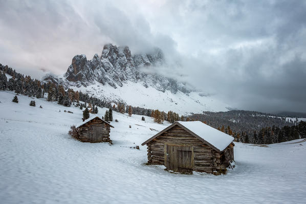 Winter scenery with two wooden lodges and Odle Dolomites covered in snow. Malga Gampen, Funes, Bolzano, Trentino Alto Adige - Sudtirol, Italy, Europe.