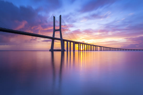 A colorful sunrise behind the Vasco da Gama bridge, reflected on the Tagus River. Lisbon, Portugal, Europe.