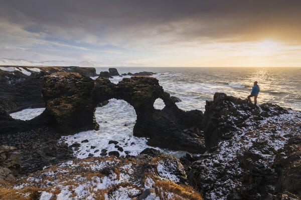 View of a man admiring the Gatklettur rock arch at sunrise in winter. Arnarstapi, Hellnar, Snæfellsnes Peninsula, Iceland, Northern Europe.