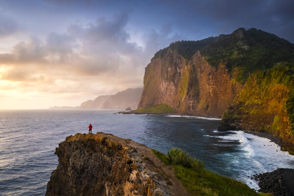 Aerial view of a tourist admiring sunrise from the cliffs of Miradouro do Guindaste. Madeira, Portugal, Europe.