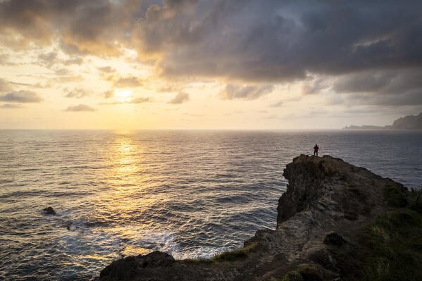 Aerial view of a tourist admiring sunrise from the cliffs of Miradouro do Guindaste. Madeira, Portugal, Europe.