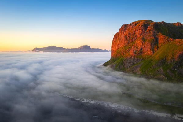Aerial view of low clouds moving around a mountain and over the town of Eggum at midnight sun in summer. Eggum Unstad, Vestvagoy Lofoten, Islands, Norway, Europe.