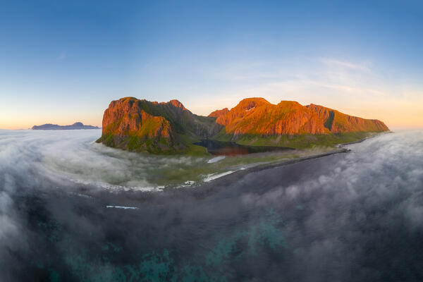 Aerial view of low clouds moving around a mountain and over the town of Eggum at midnight sun in summer. Eggum Unstad, Vestvagoy Lofoten, Islands, Norway, Europe.