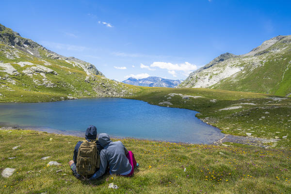 Rear view of man and woman in love sitting on grass admiring Baldiscio lakes, Valchiavenna, Vallespluga, Lombardy, Italy