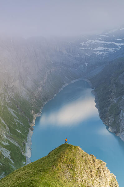 Aerial view of hiker man on rocks looking at lake Limmernsee in the mist at sunset, Canton of Glarus, Switzerland